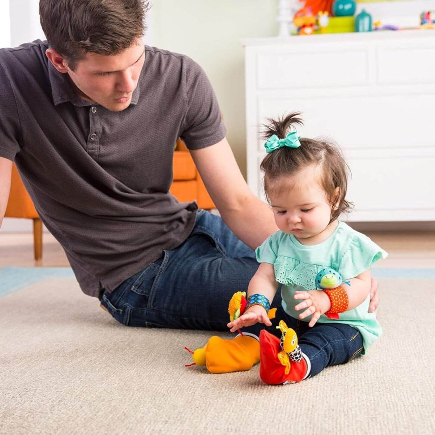 A baby sitting with a parent, playing with the Lamaze Gardenbug Foot Finder and Wrist Rattle Set. Colourful bugs encourage sensory play and movement.