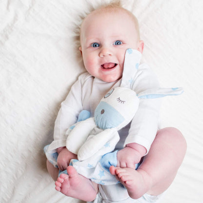 Smiling baby with bright blue eyes holding the Lulujo Blue Puppy Muslin Lovey security blanket against white bedding, showing bond with comfort toy.