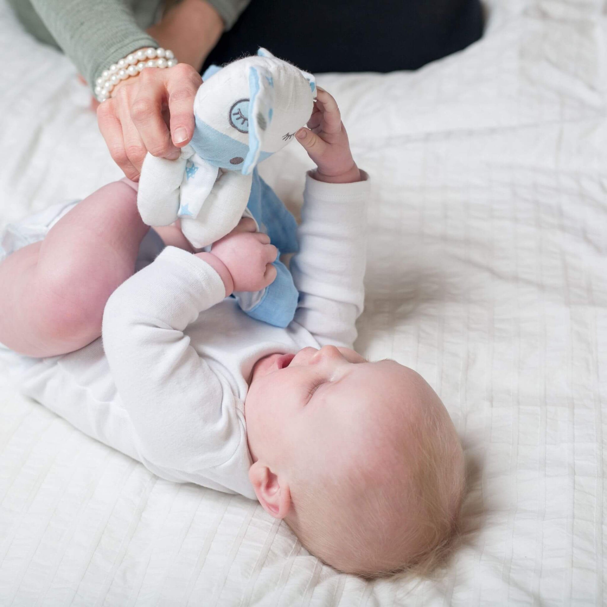 Peaceful baby lying on white bedding while parent introduces the Lulujo Blue Puppy Muslin Lovey, perfect first comfort companion.