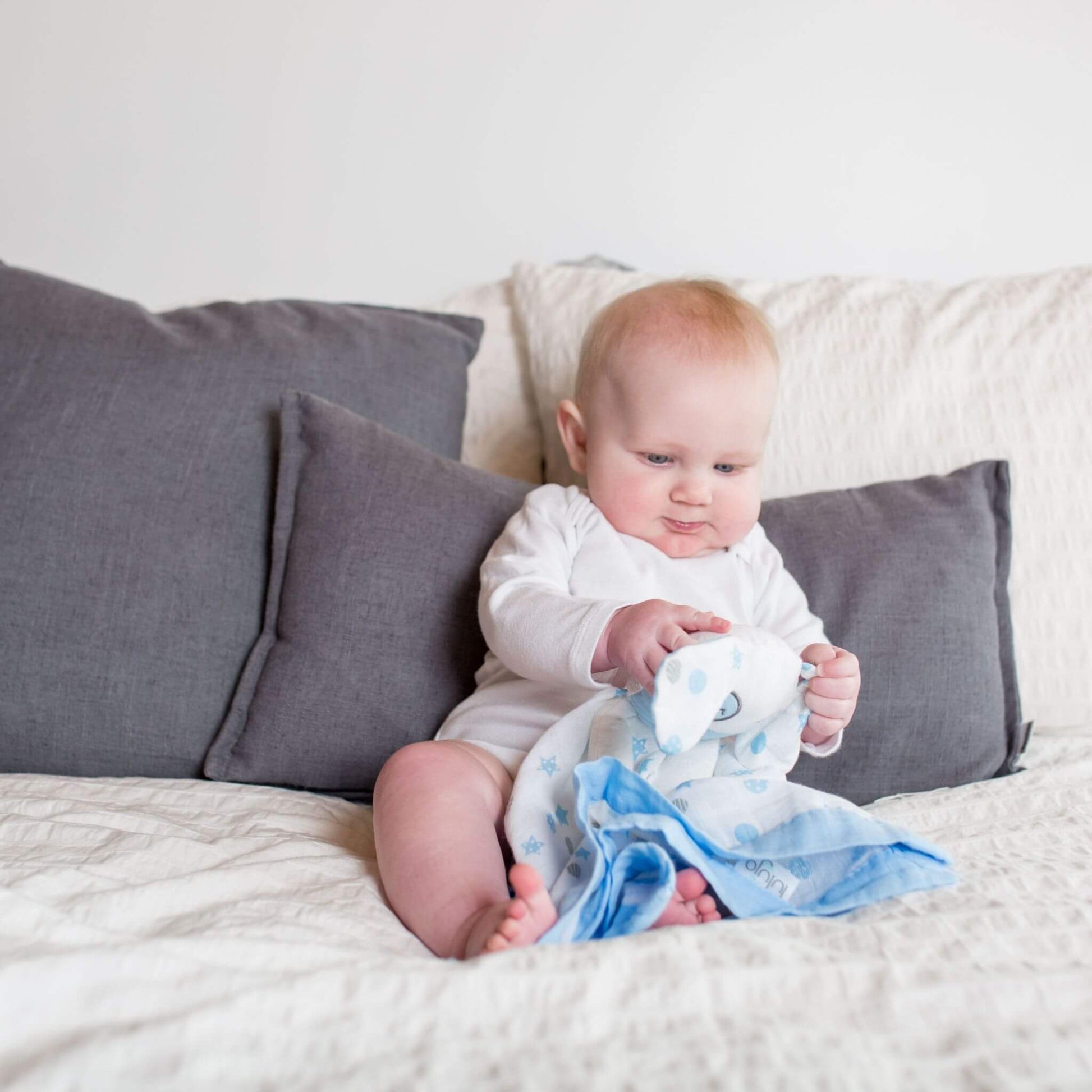 Baby sitting on couch examining the Lulujo Blue Puppy Muslin Lovey, showing how the comforter's soft texture and puppy design captivate little ones.