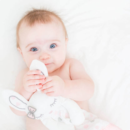 Baby with bright blue eyes holding the Lulujo Pink Bunny Lovey security blanket against white bedding, demonstrating bonding with comfort toy.