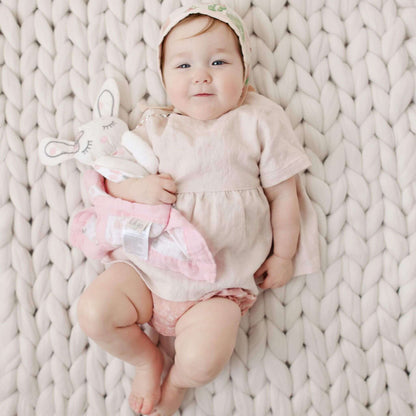 Infant in light beige outfit lying on chunky knit blanket cuddling with Lulujo Pink Bunny Lovey, showing perfect size for small hands.