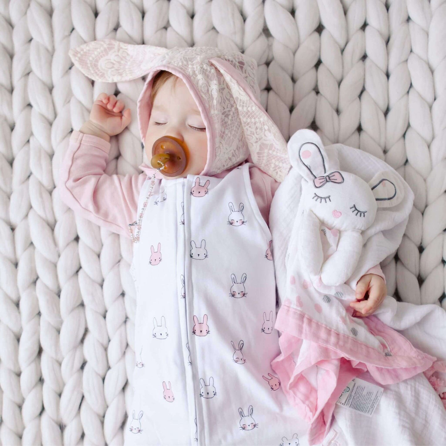 Sleeping baby with pacifier wearing bunny-themed sleepsuit and bunny bonnet while holding Lulujo Pink Bunny Lovey on chunky knit blanket.