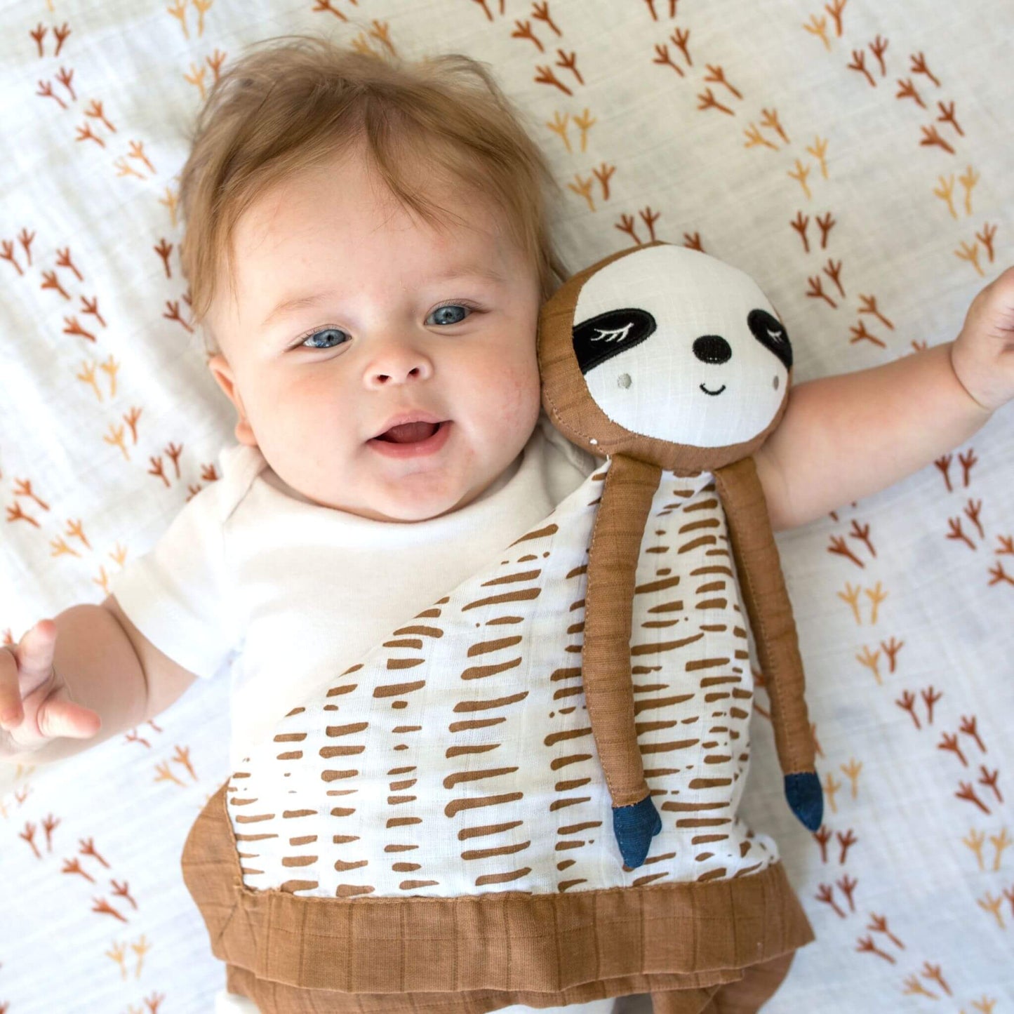 Happy baby with blue eyes cuddling with Lulujo Sloth Muslin Lovey on patterned bedding, showcasing the perfect size of this adorable comforter.