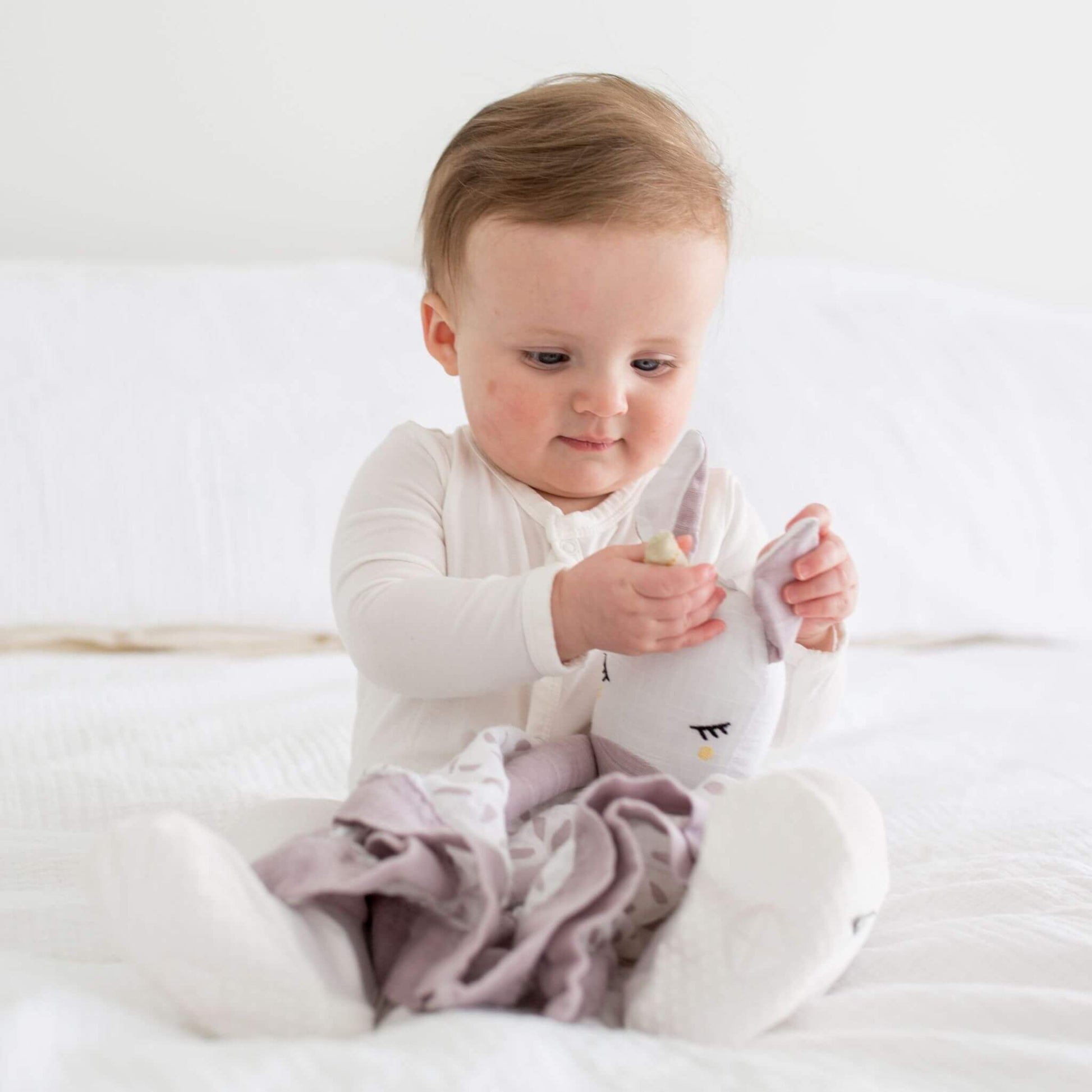 Focused baby examining their Lulujo Unicorn Muslin Lovey on white bedding, showing how the lovey's soft texture captivates little hands.