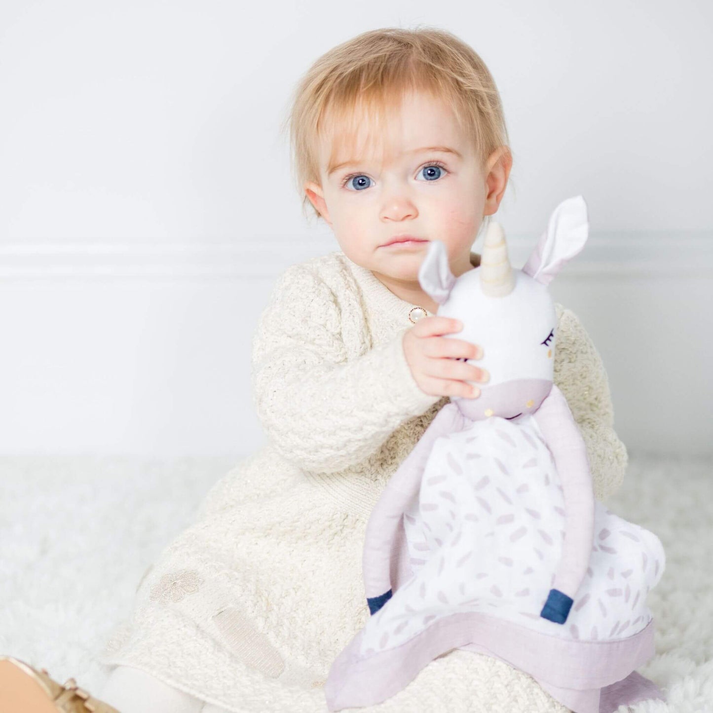 Toddler with bright blue eyes holding the Lulujo Unicorn Muslin Lovey against cream knit clothing, highlighting the lovey's perfect size for small hands.