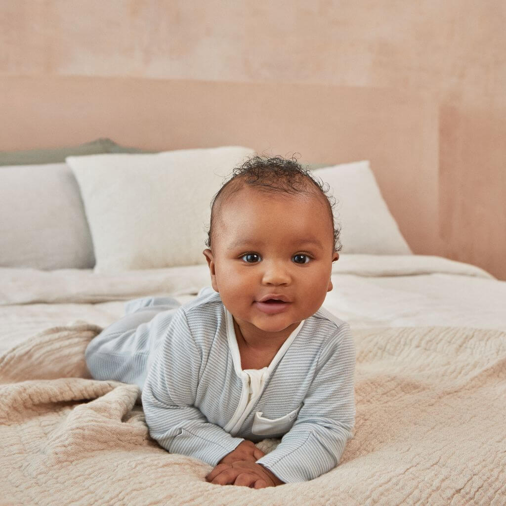 Baby in the MORI Clever Zip Sleepsuit, blue stripe, on a bed. The sleepsuit features a front zip and small pocket, designed for comfort and easy changing.