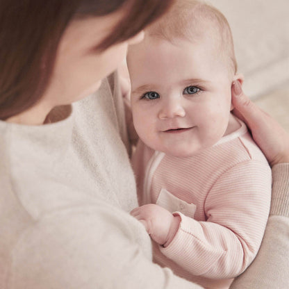 Baby wearing the MORI Clever Zip Sleepsuit in blush stripe, smiling while being held by their mother.