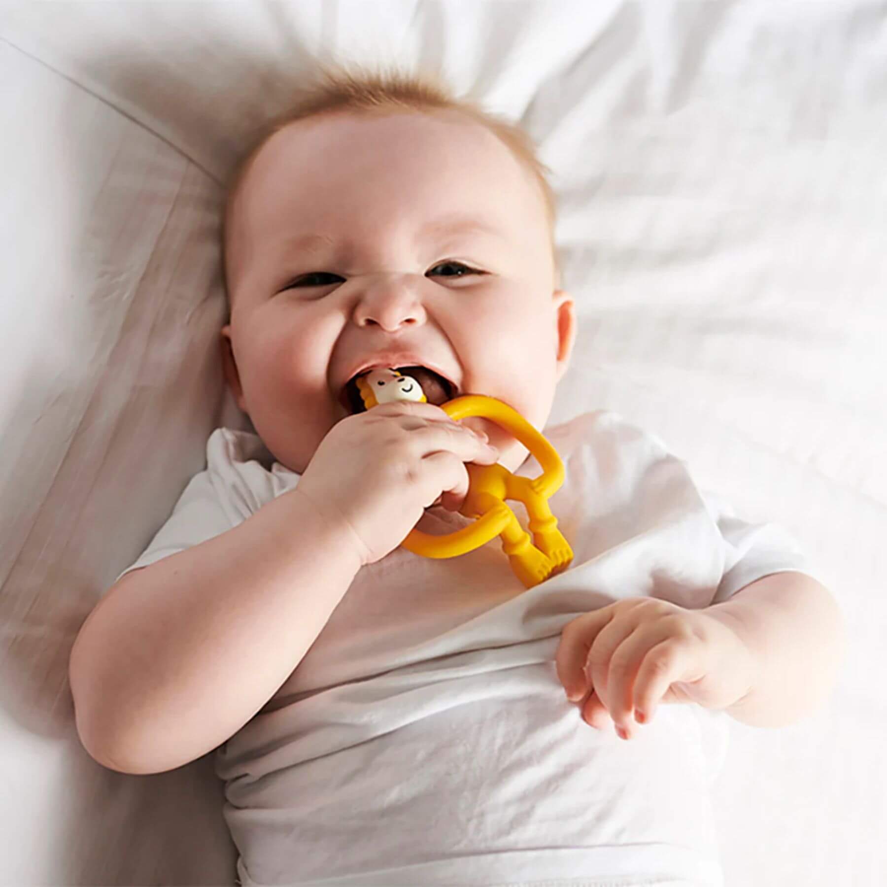 A baby lying on a bed, smiling and holding a yellow Matchstick Monkey Ludo Lion teether. The baby is using the teether for comfort, wearing a white outfit.