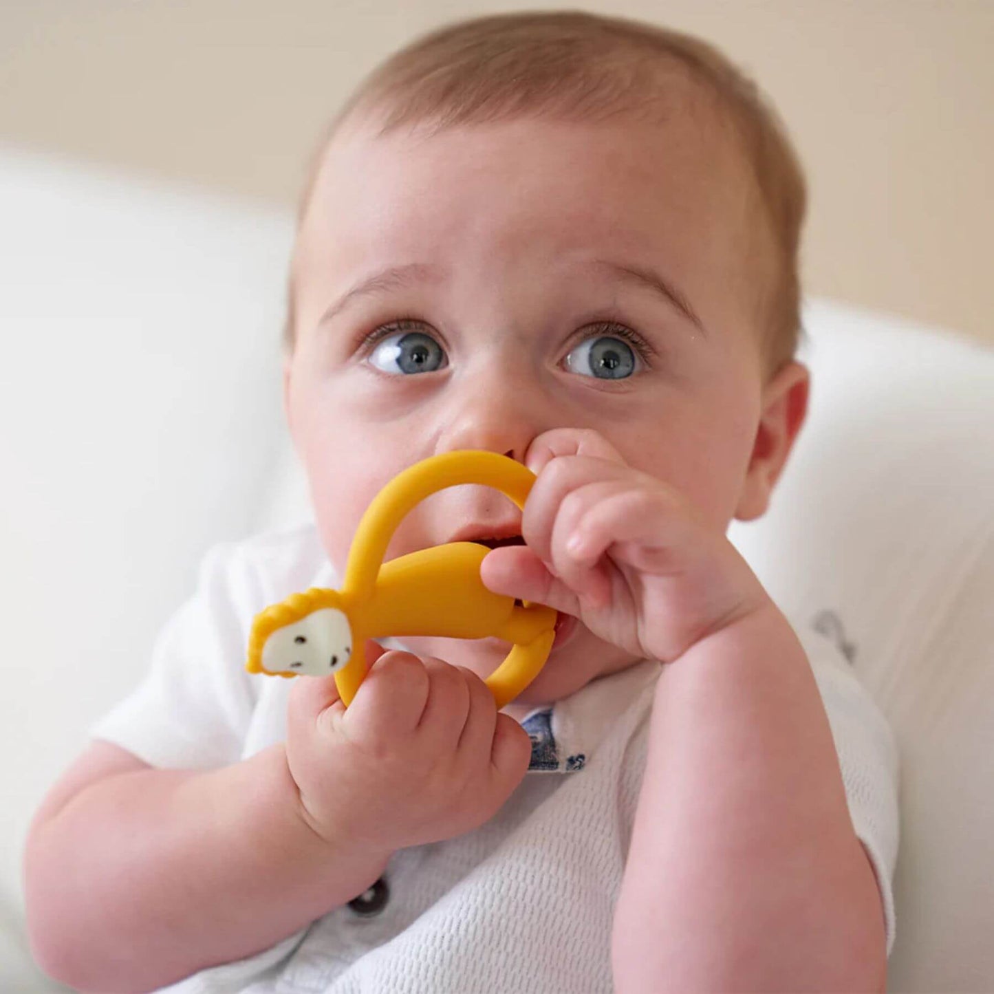 A baby holding and chewing on a yellow Matchstick Monkey Ludo Lion teether. The baby is seated, wearing a white outfit and using the teether for teething relief.