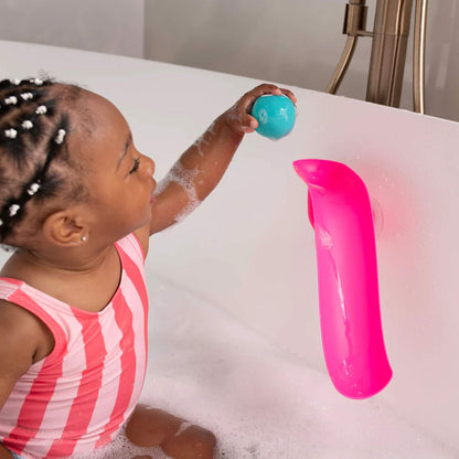 A toddler in a striped swimsuit playing in a soapy bath, holding a teal animal rock over a pink Matchstick Monkey slide attached to the tub wall.