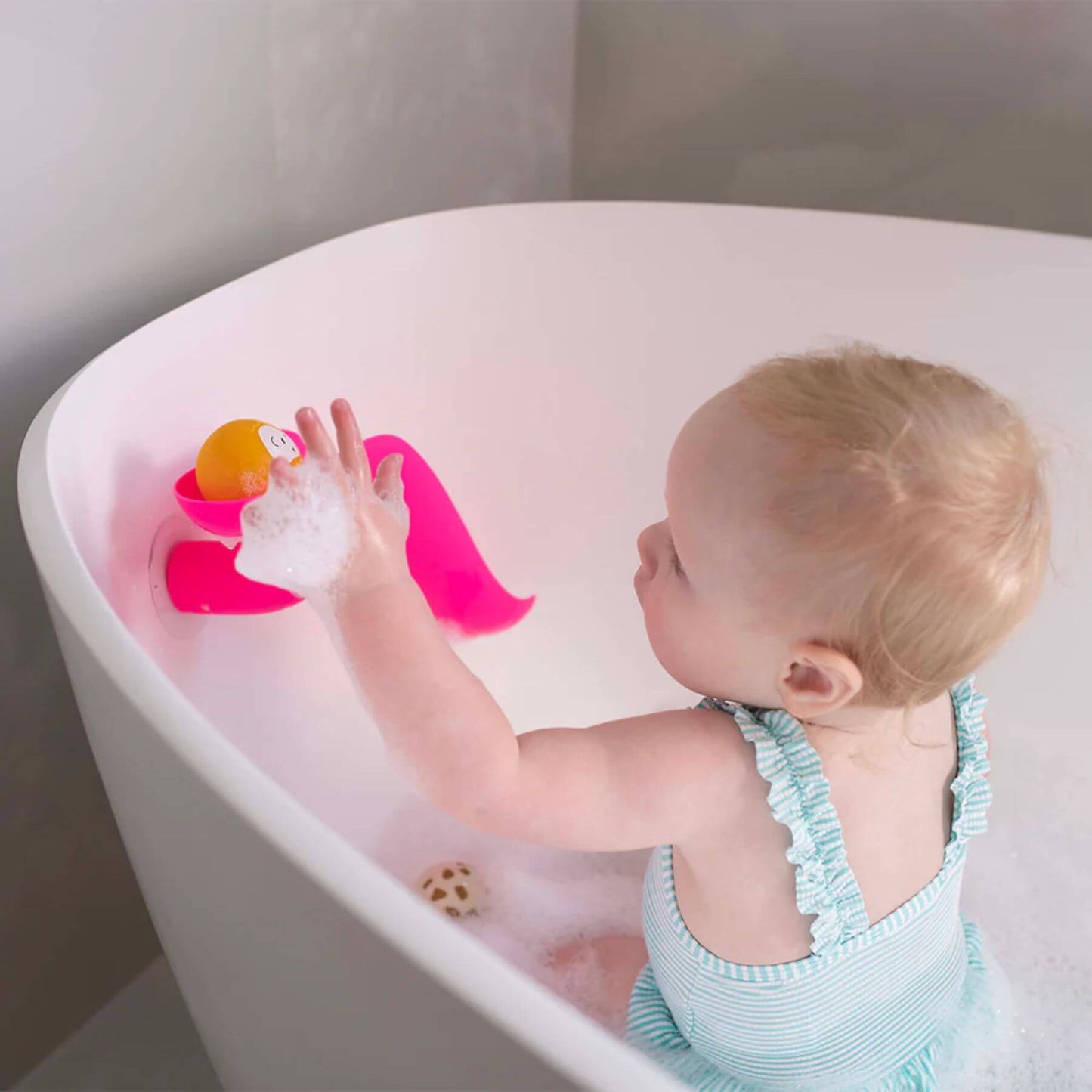 A toddler in a striped swimsuit playing in a soapy bath, using a pink Matchstick Monkey slide to drop a yellow lion animal rock down into the water.