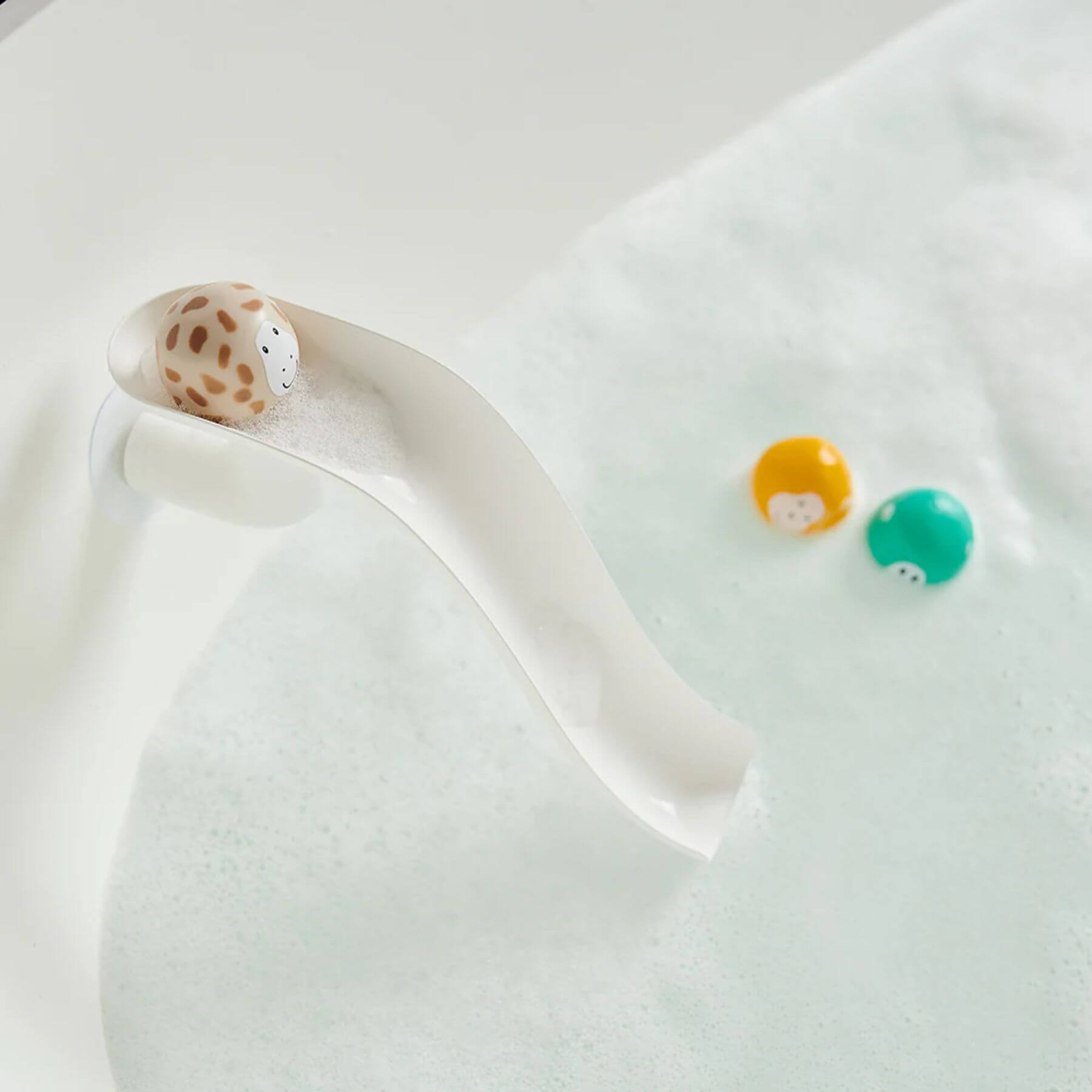 A white Matchstick Monkey slide attached to a bath, with a giraffe animal rock at the top and a yellow lion and teal monkey floating in soapy water below