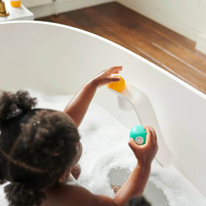 A toddler in a soapy bath playing with a white Matchstick Monkey slide, holding a yellow lion animal rock at the top and a teal monkey animal rock in the other hand. 