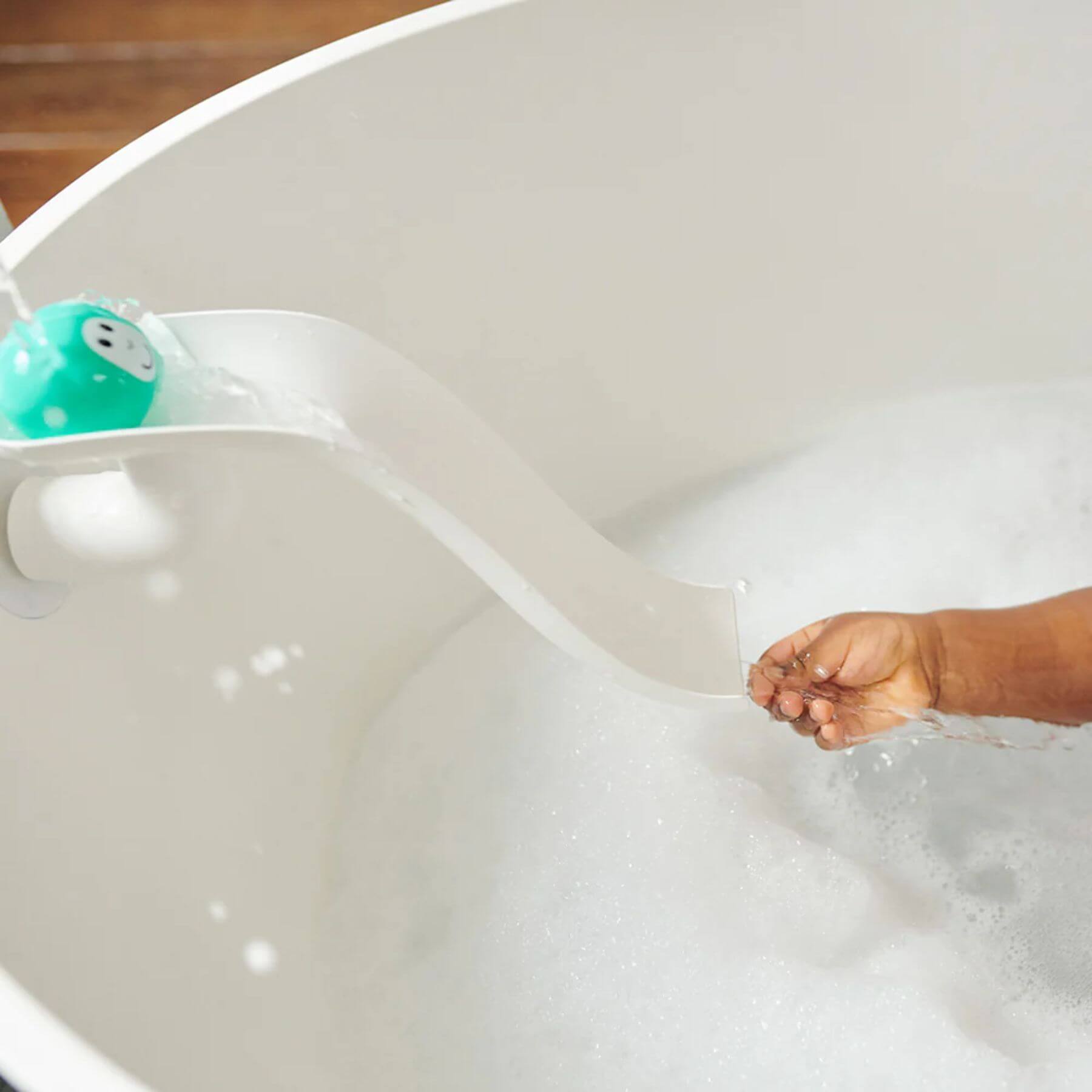 A child’s hand reaching out to catch water as a teal Matchstick Monkey animal rock slides down a white bath slide into soapy water below.