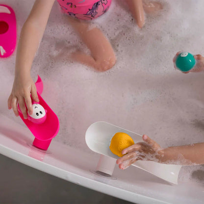 Two children in a soapy bath playing with pink and white Matchstick Monkey slides. Each child is placing colourful animal rocks on the slides.