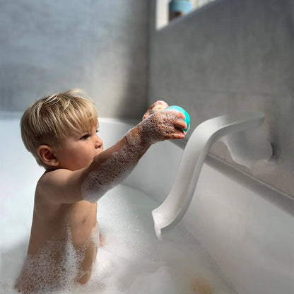 A toddler in a soapy bath reaching up to place a teal and beige animal animal rock onto a white Matchstick Monkey slide attached to the bathtub.