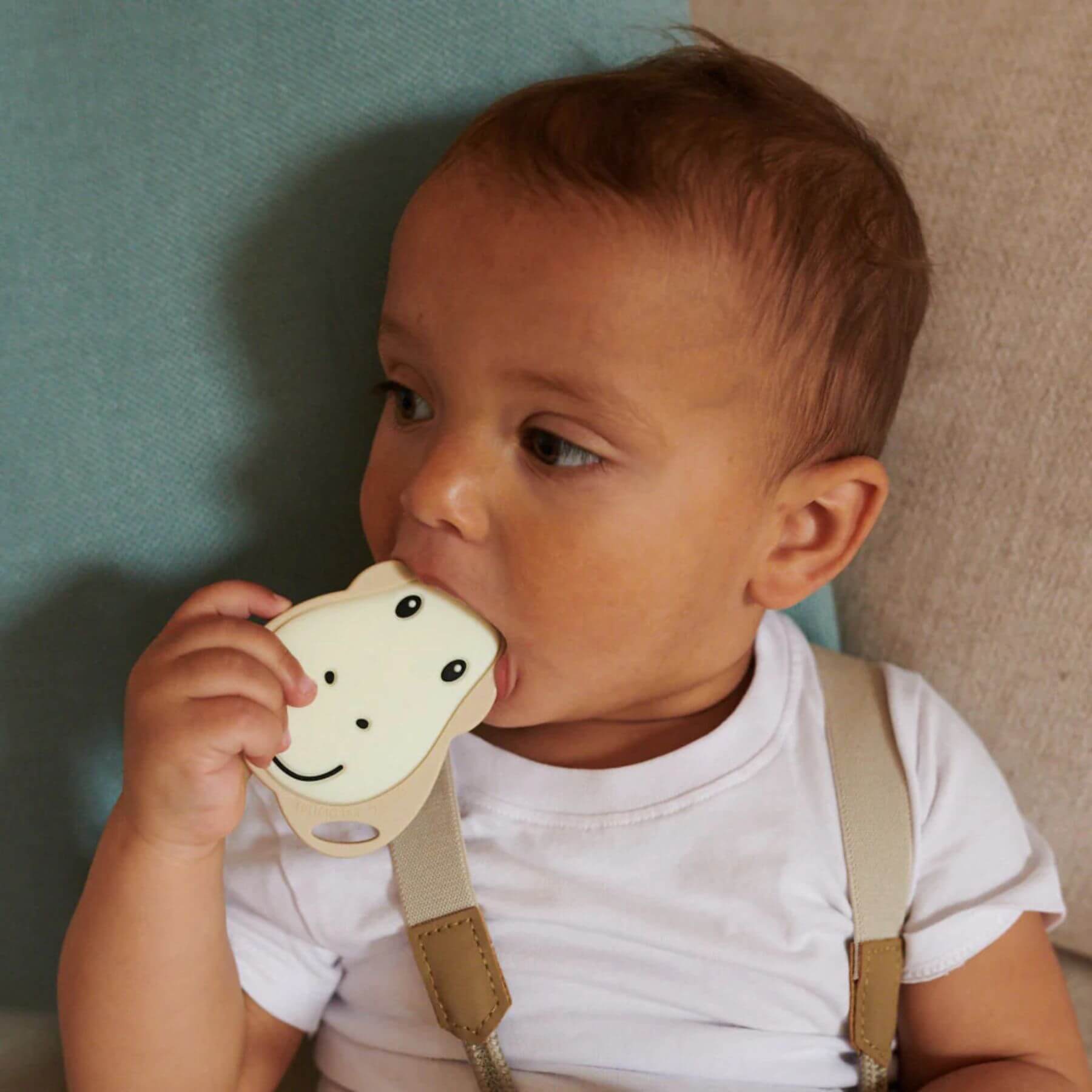 A baby chewing on a beige Matchstick Monkey giraffe teether while sitting on a sofa. The teether has a smiling giraffe face, designed for teething relief.