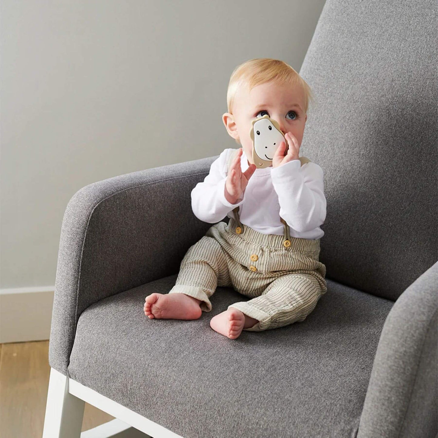 A baby sitting on a grey chair, holding a beige Matchstick Monkey giraffe teether to their mouth. The baby is wearing a white top and beige trousers.