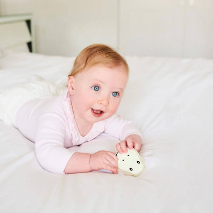 A baby lying on a bed, smiling and holding a beige Matchstick Monkey giraffe teether, wearing a pink outfit, with a playful expression