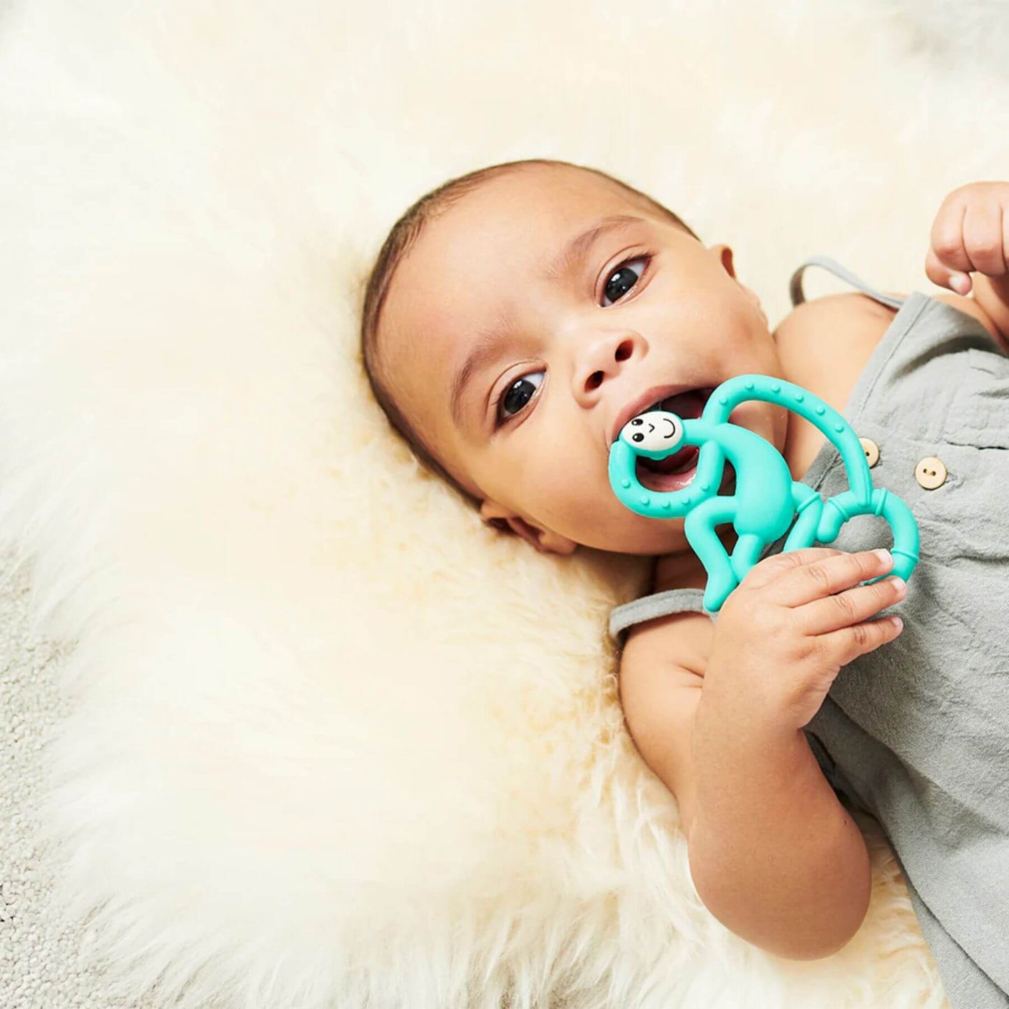 Baby holding a green Matchstick Monkey mini teether while lying down. The teether is designed for soothing gums with textured silicone.