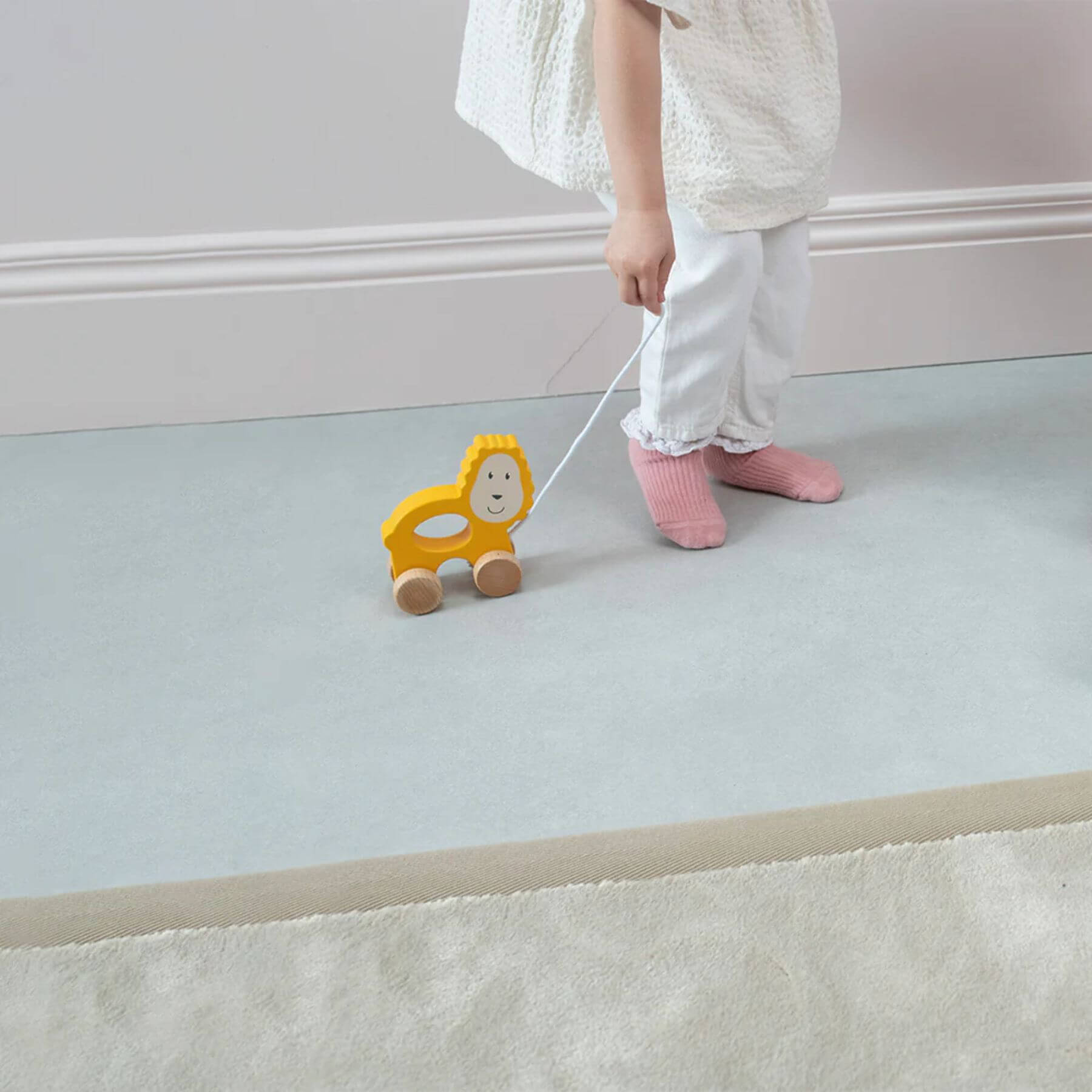 A child wearing pink socks and white trousers pulls a matchstick monkey wooden lion toy along the floor by its string, standing next to a soft rug.