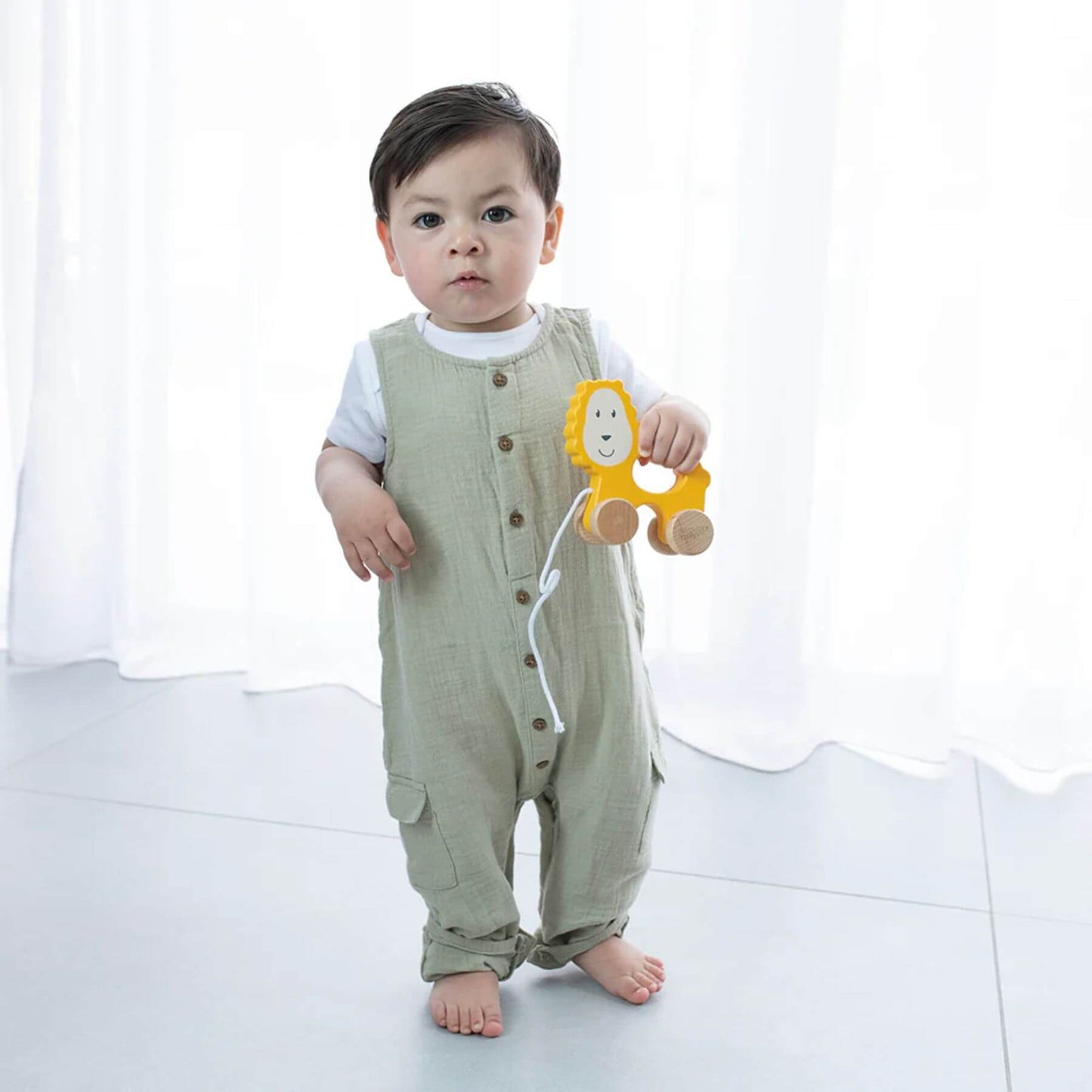 A toddler stands barefoot holding a Matchstick Monkey wooden pull-along lion toy, wearing a green romper with soft light filtering through white curtains.