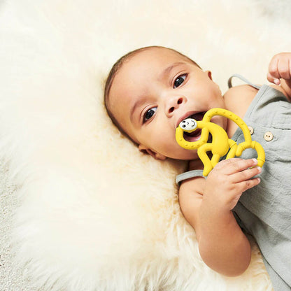 A baby lies on a soft fluffy surface while holding a yellow Matchstick Monkey mini teether. The baby is chewing on the textured teether, designed to soothe gums.