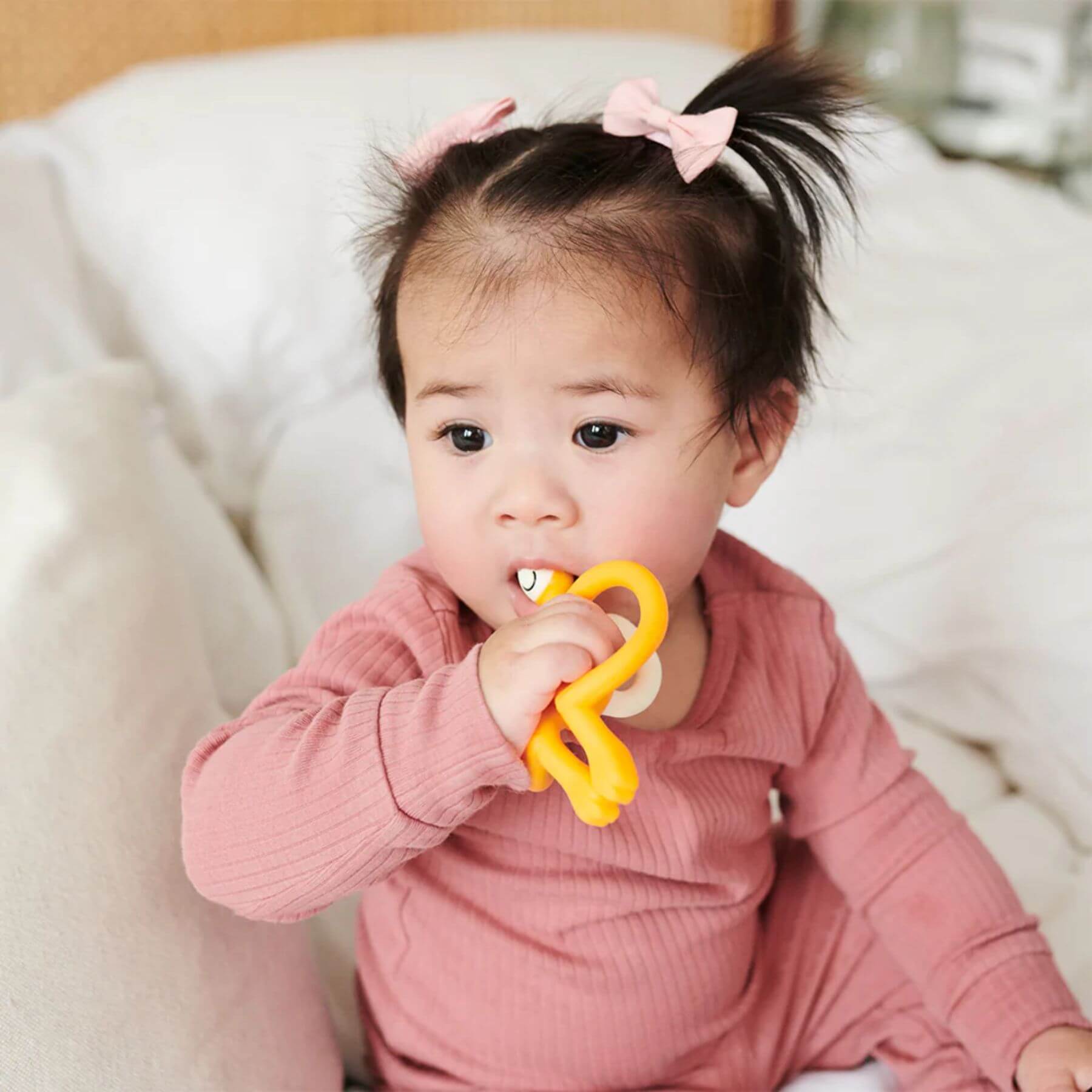 
A baby is sitting on a bed, holding and chewing on a bright yellow Matchstick Monkey Original Teether toy. The baby wears a pink outfit with cute bows in her hair.