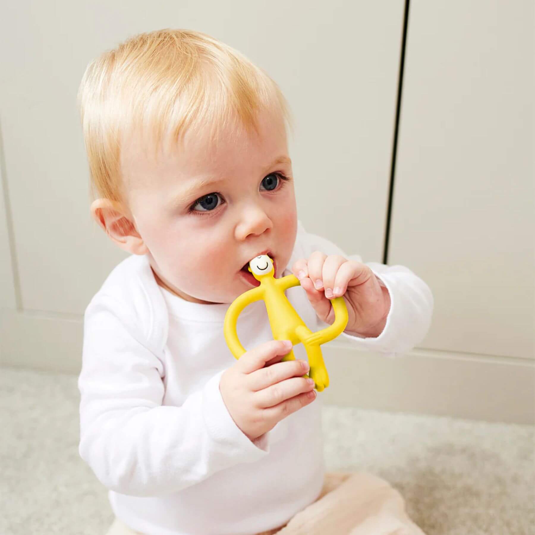 A baby is shown sitting and holding a Matchstick Monkey original teether toy in yellow. The baby is biting the teether with both hands, helping to soothe their gums. The teether’s design makes it easy for the baby to grip.