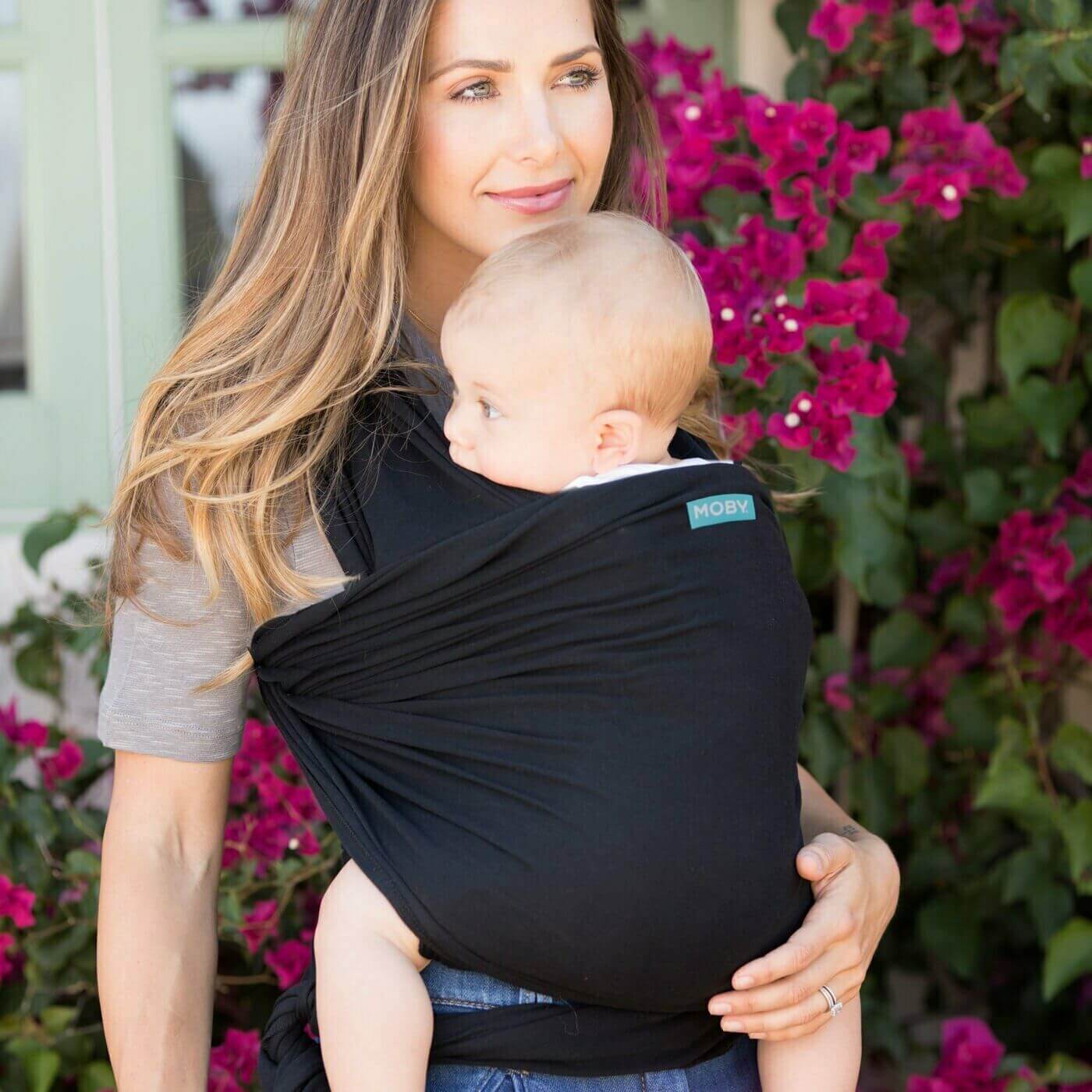 A mother wearing a baby in a black Classic Moby wrap, standing in front of a vibrant background of blooming pink flowers.