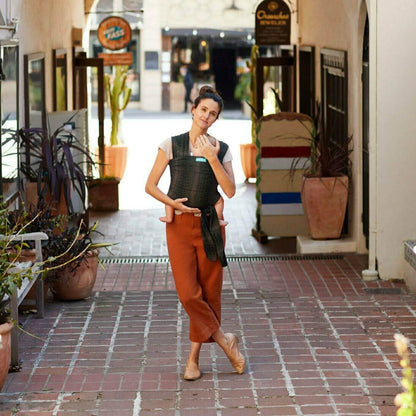 Mother wearing a Moby Fleck baby wrap, walking through a charming outdoor corridor with potted plants and storefronts in the background.