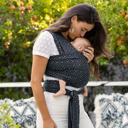 Mother kissing her baby while carrying them in a Moby Mosaic wrap, enjoying a secure and stylish babywearing experience outdoors with a lush green garden and fruit trees in the background.