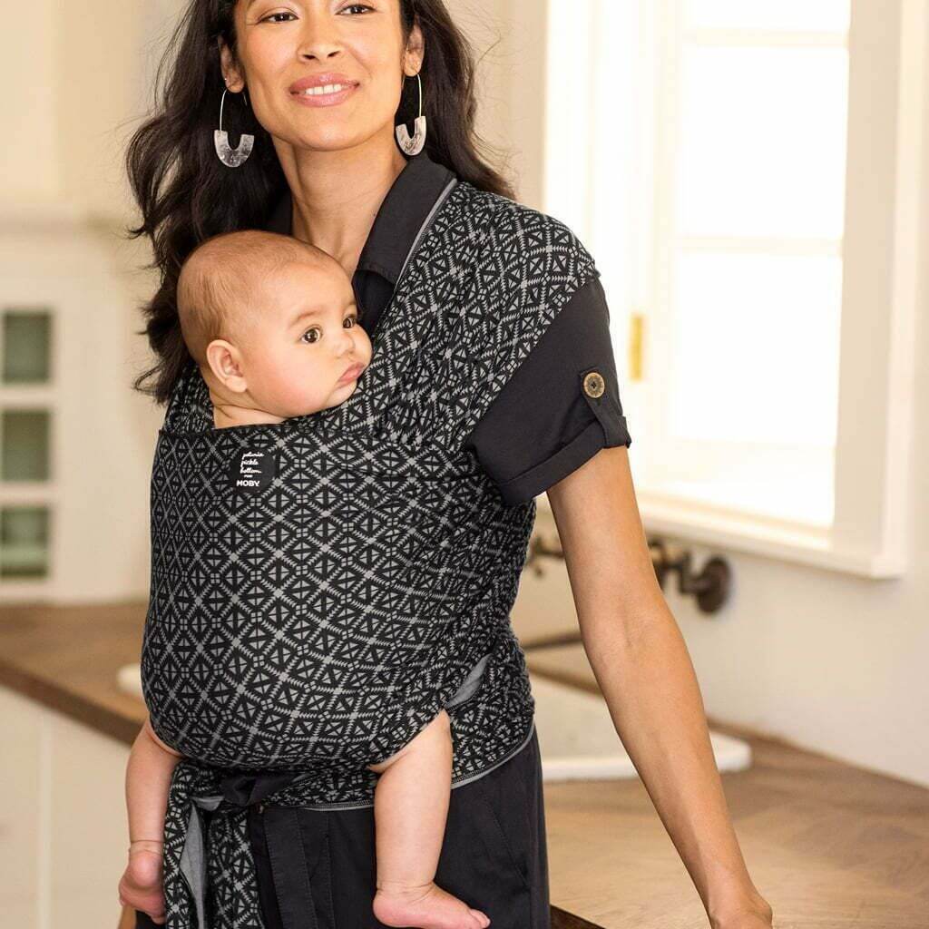 Mother wearing a Moby Mosaic baby wrap, holding her baby in a cosy indoor kitchen setting with warm lighting and wooden countertops in the background.