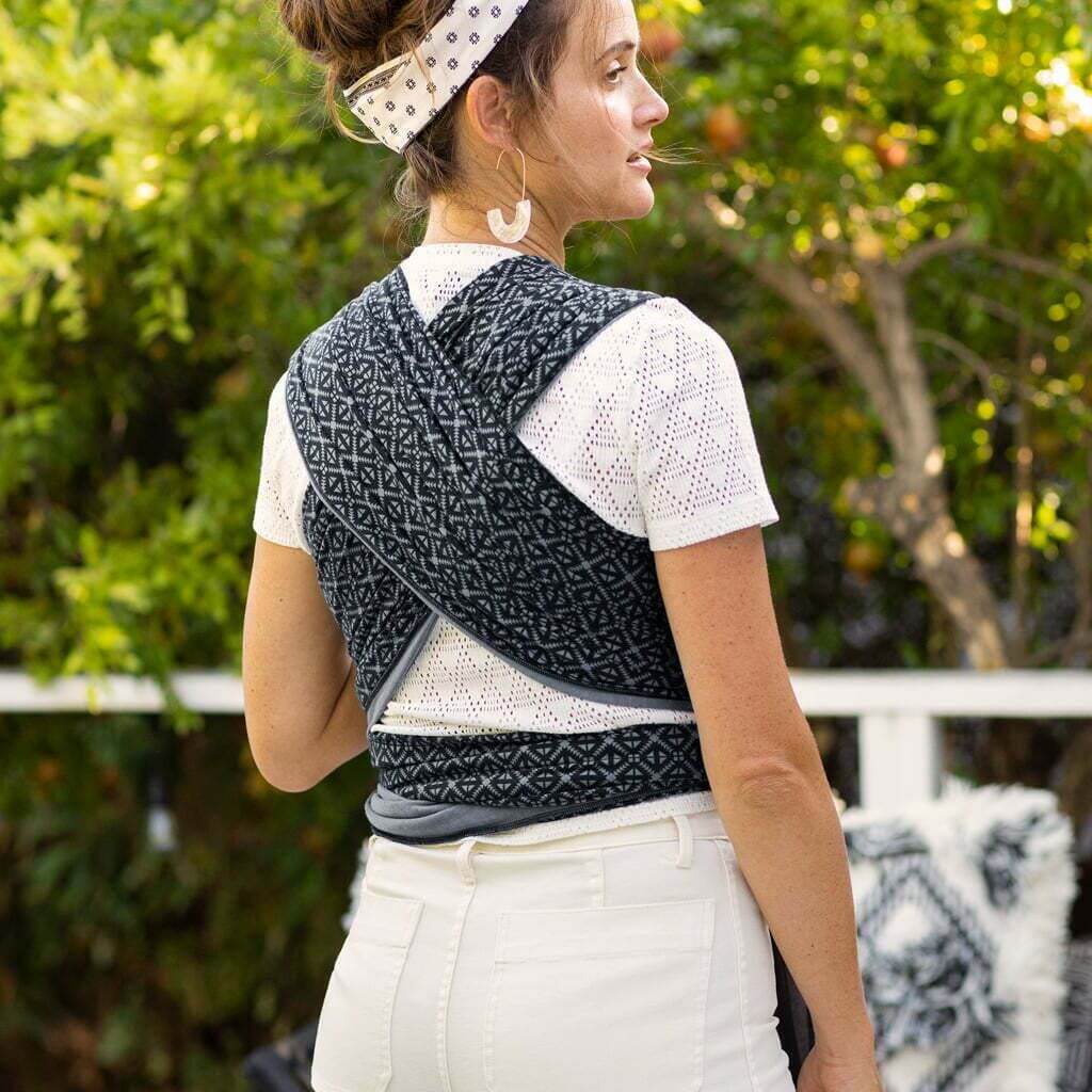 Rear view of a mother wearing a Moby Mosaic baby wrap with a criss-cross back design, standing outdoors with lush greenery and trees in the background.