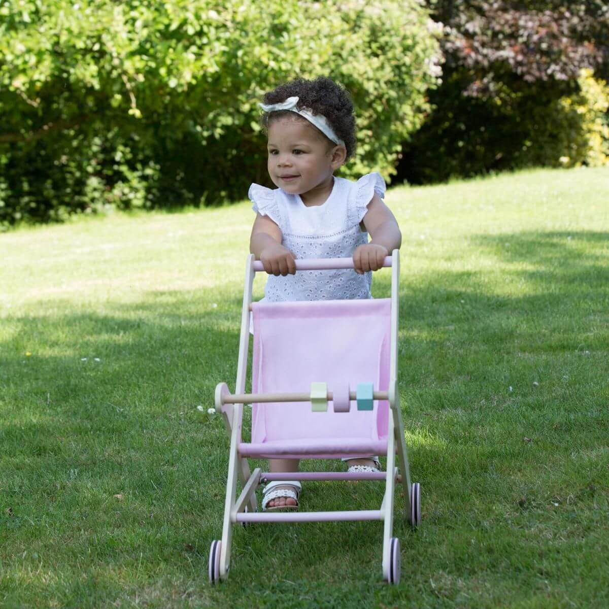 Toddler pushing a Moover Toys pink wooden dolls' stroller with pastel-coloured beads and natural wood frame, enjoying outdoor play while developing motor skills and coordination.