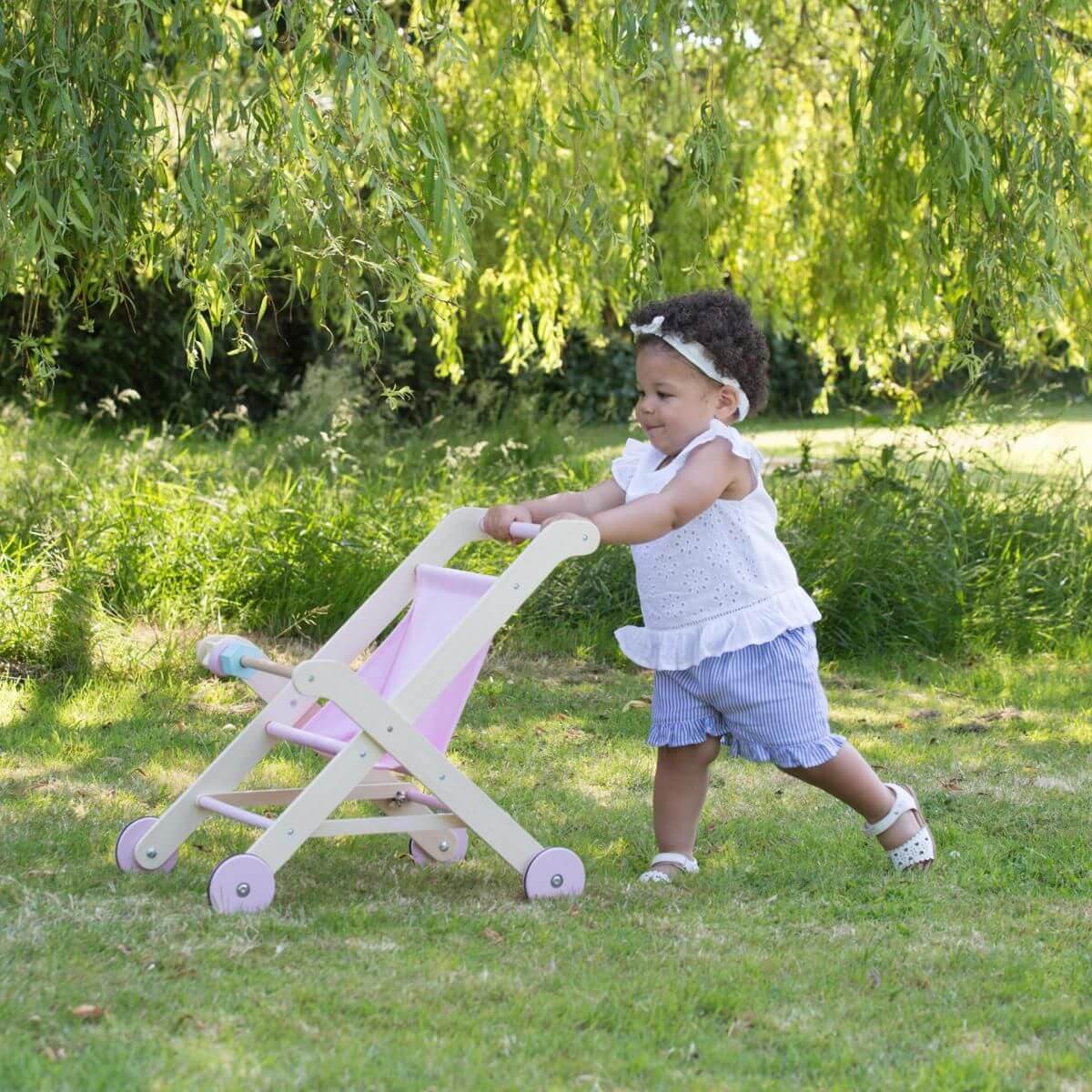 Toddler pushing a Moover Toys pink wooden dolls' stroller outdoors. The child is walking confidently on a grassy lawn, wearing a white sleeveless top, striped shorts, and sandals.