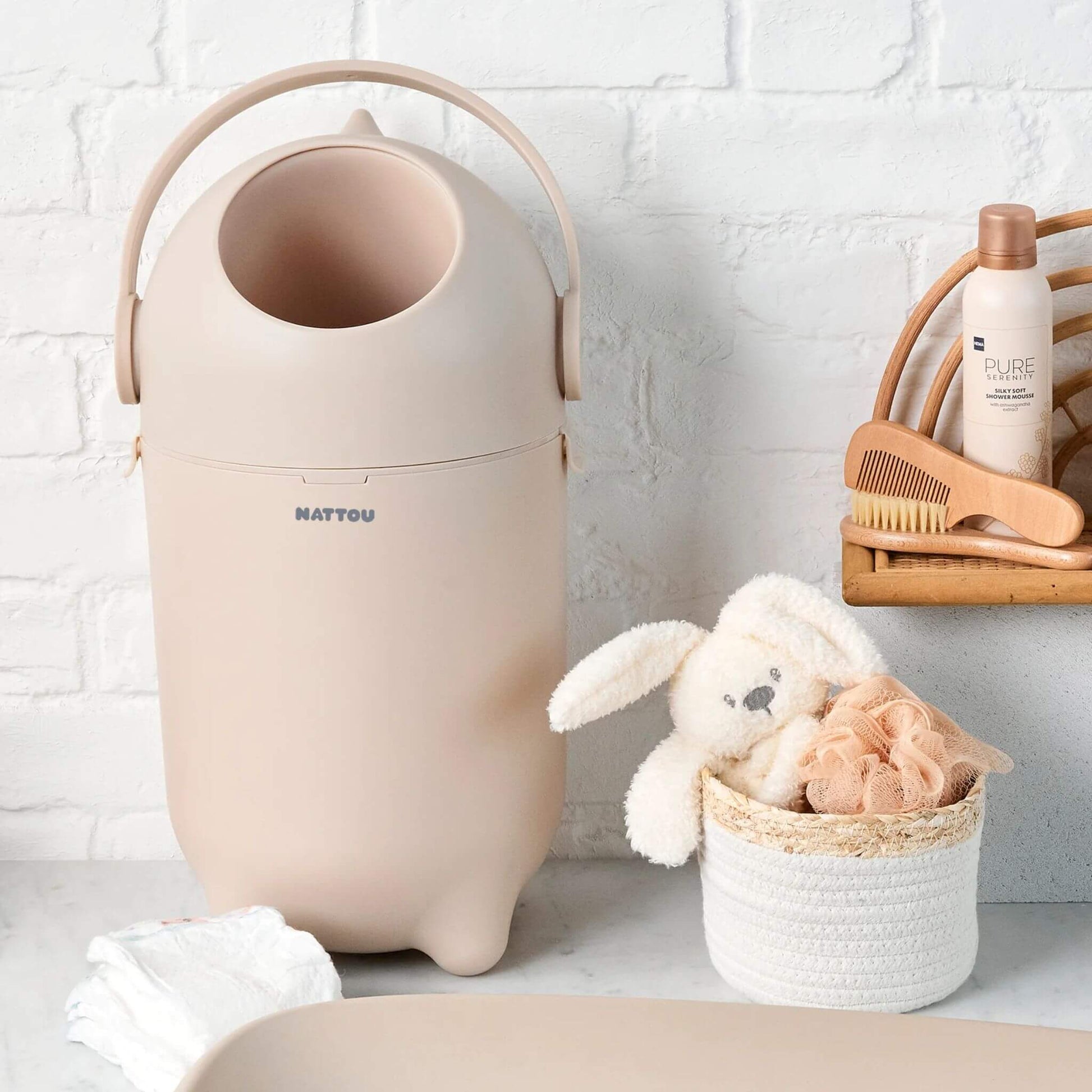 The Nattou Dropy Nappy Bin placed in a nursery setting, next to a basket with a plush bunny and baby essentials.