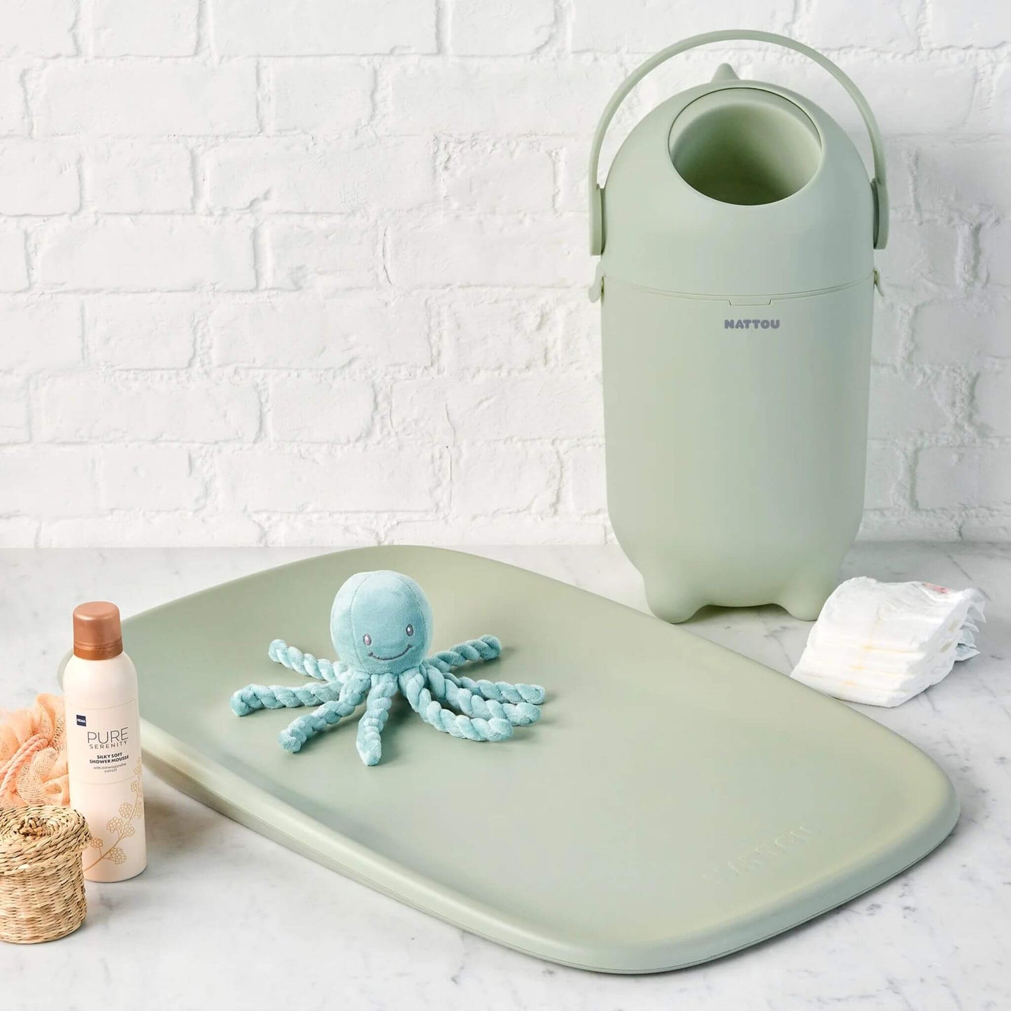 The green Nattou Dropy Nappy Bin in a nursery setup, placed next to a matching changing mat and baby essentials.