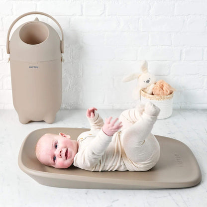 The taupe Nattou Dropy Nappy Bin in a nursery setting, placed beside a changing mat with a smiling baby lying on it.