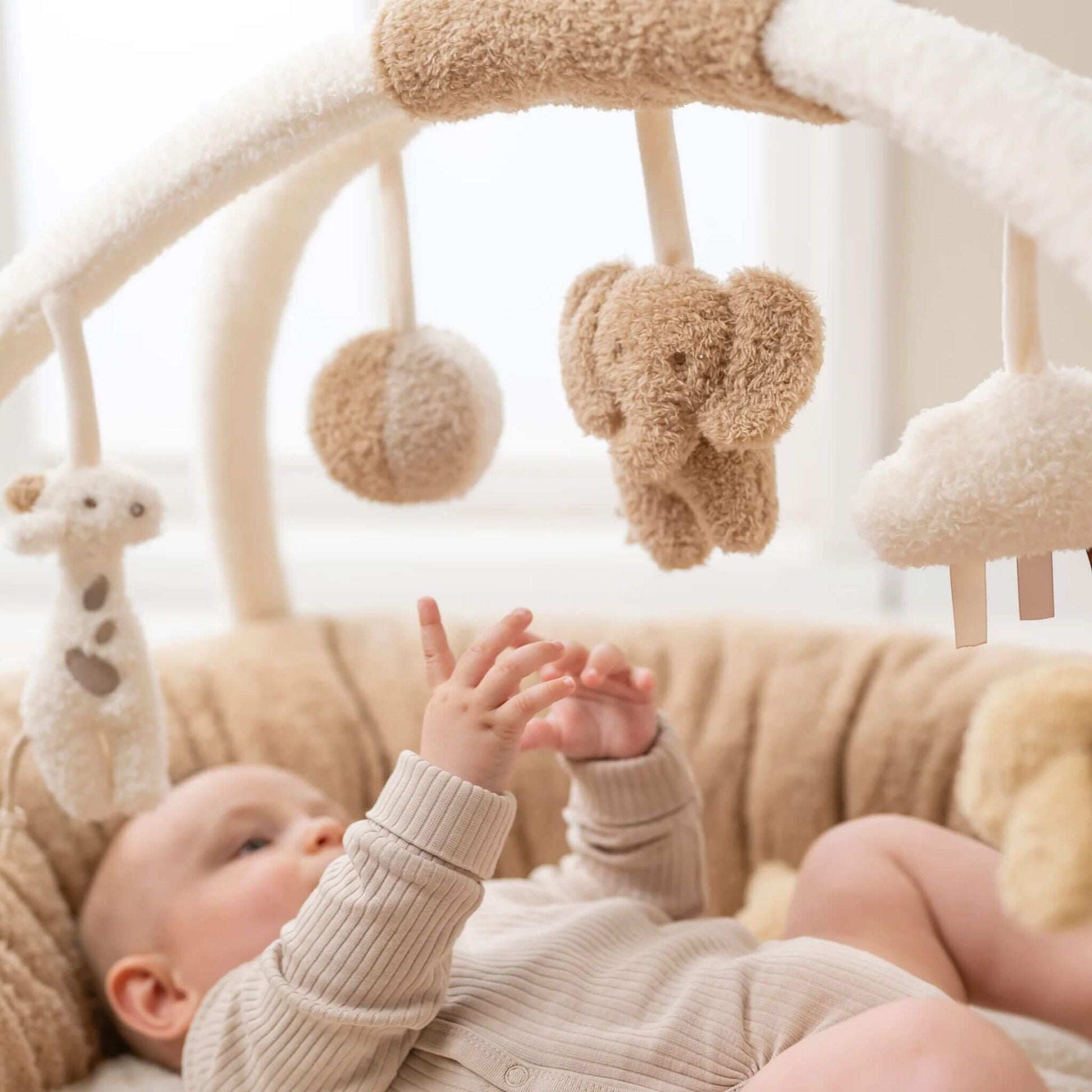 Baby reaching for plush hanging toys on the detachable arch of the Nattou Teddy Playmat, engaging with the elephant, giraffe, and cloud.