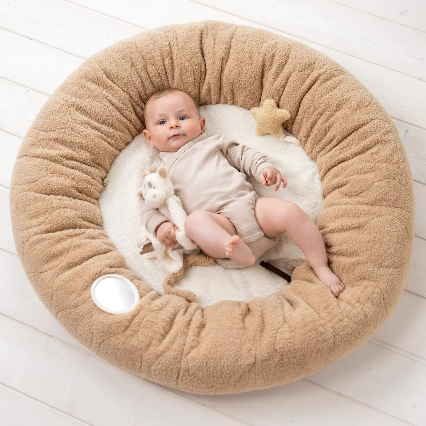 Baby lying on the Nattou Teddy Playmat, holding a plush giraffe, surrounded by soft textures, a star, textured labels, and a mirror.