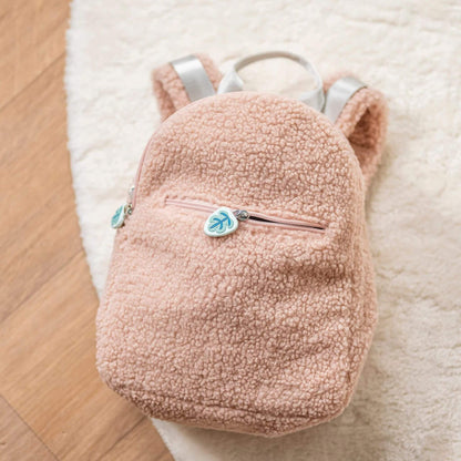 Soft pink Nattou Teddy Backpack with a plush, textured fabric, zippered front pocket, and adjustable straps, resting on a cosy rug.