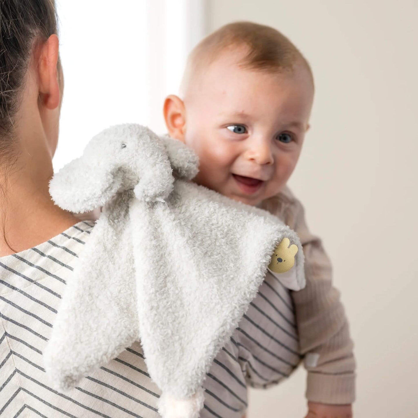 Smiling baby with Nattou Elephant Doudou comfort blanket, showing soft grey plush material and soothing design features."