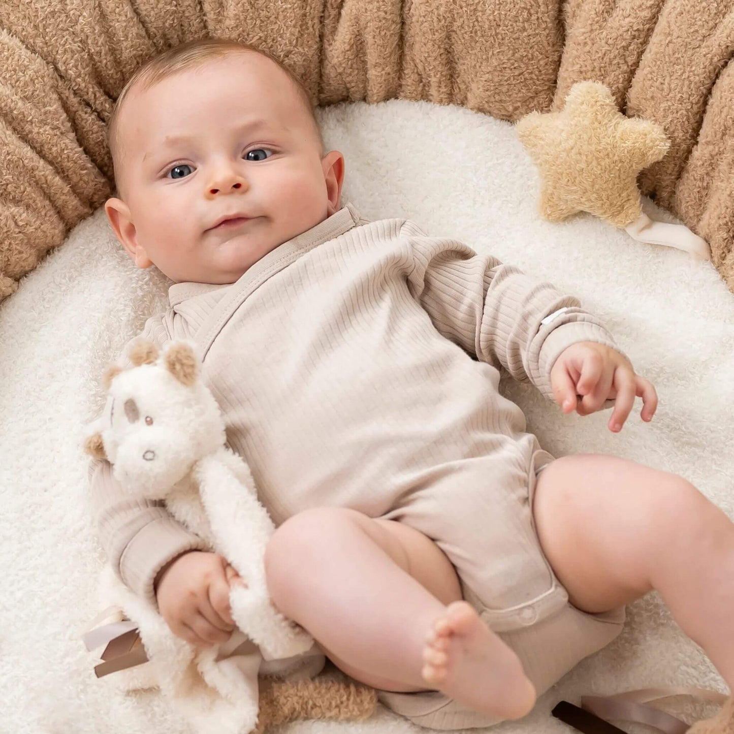 Baby in beige outfit cuddling Nattou Teddy Giraffe Doudou comfort blanket on soft white blanket.