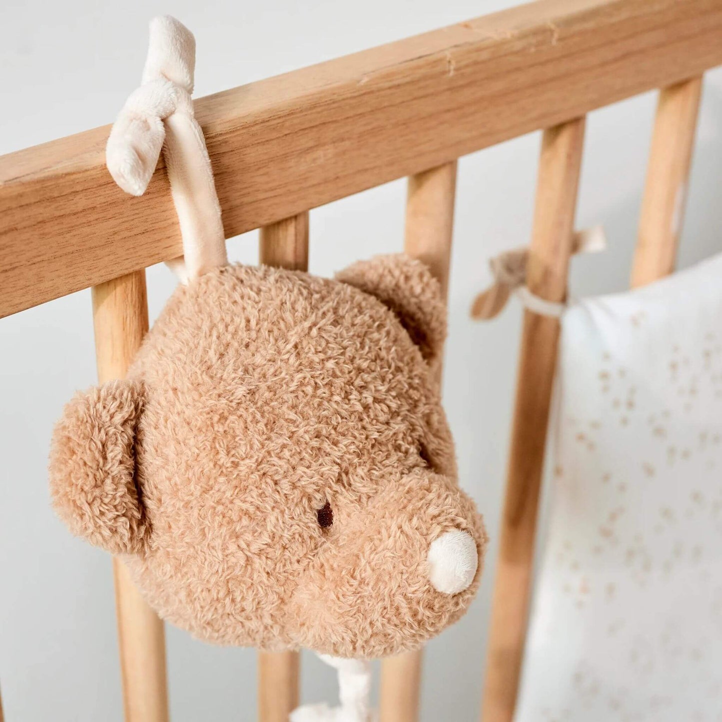 Nattou Bear teddy musical toy hanging from wooden crib rail, featuring soft beige texture and cream ribbon attachment.