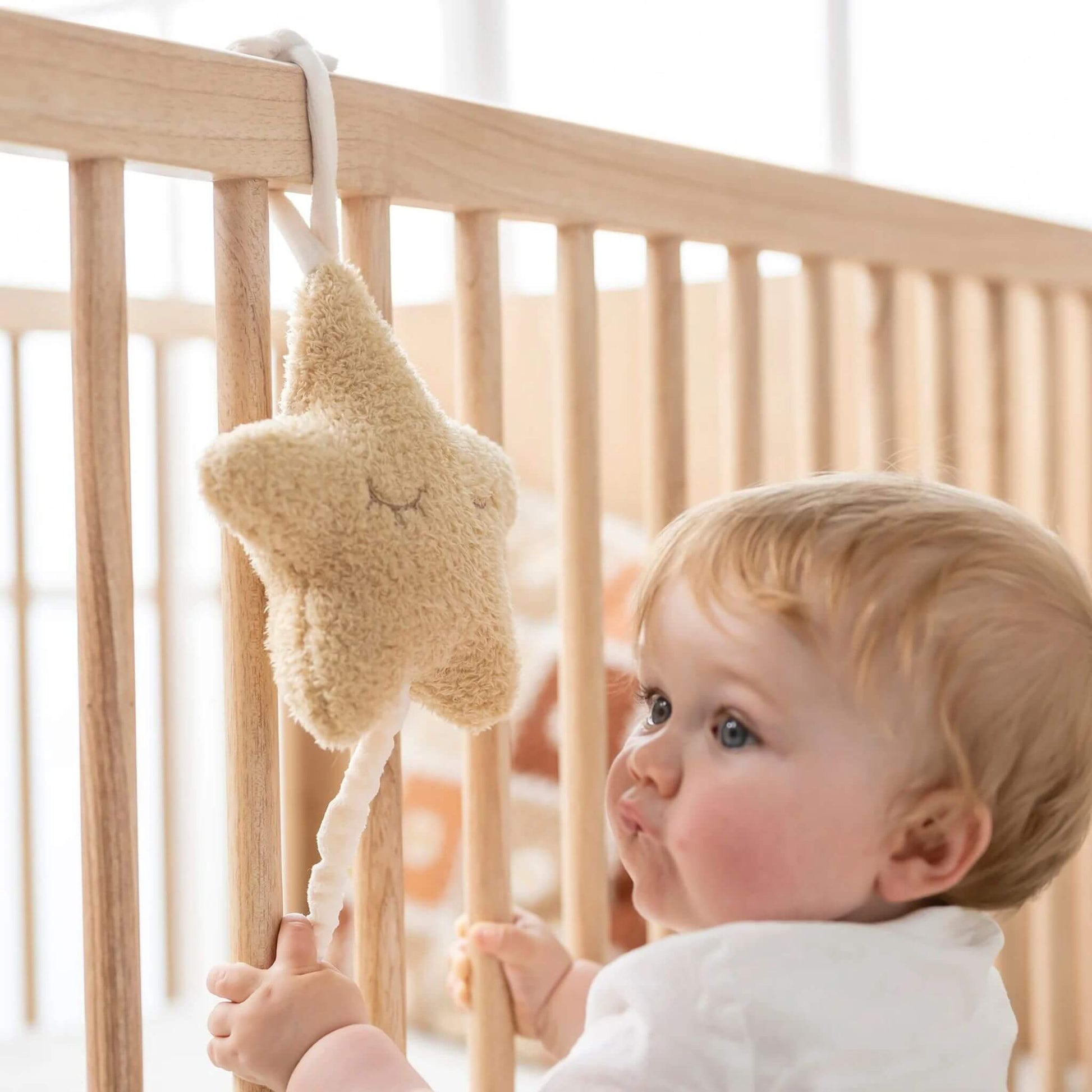 Baby standing by a wooden cot, reaching for the hanging plush star musical toy.