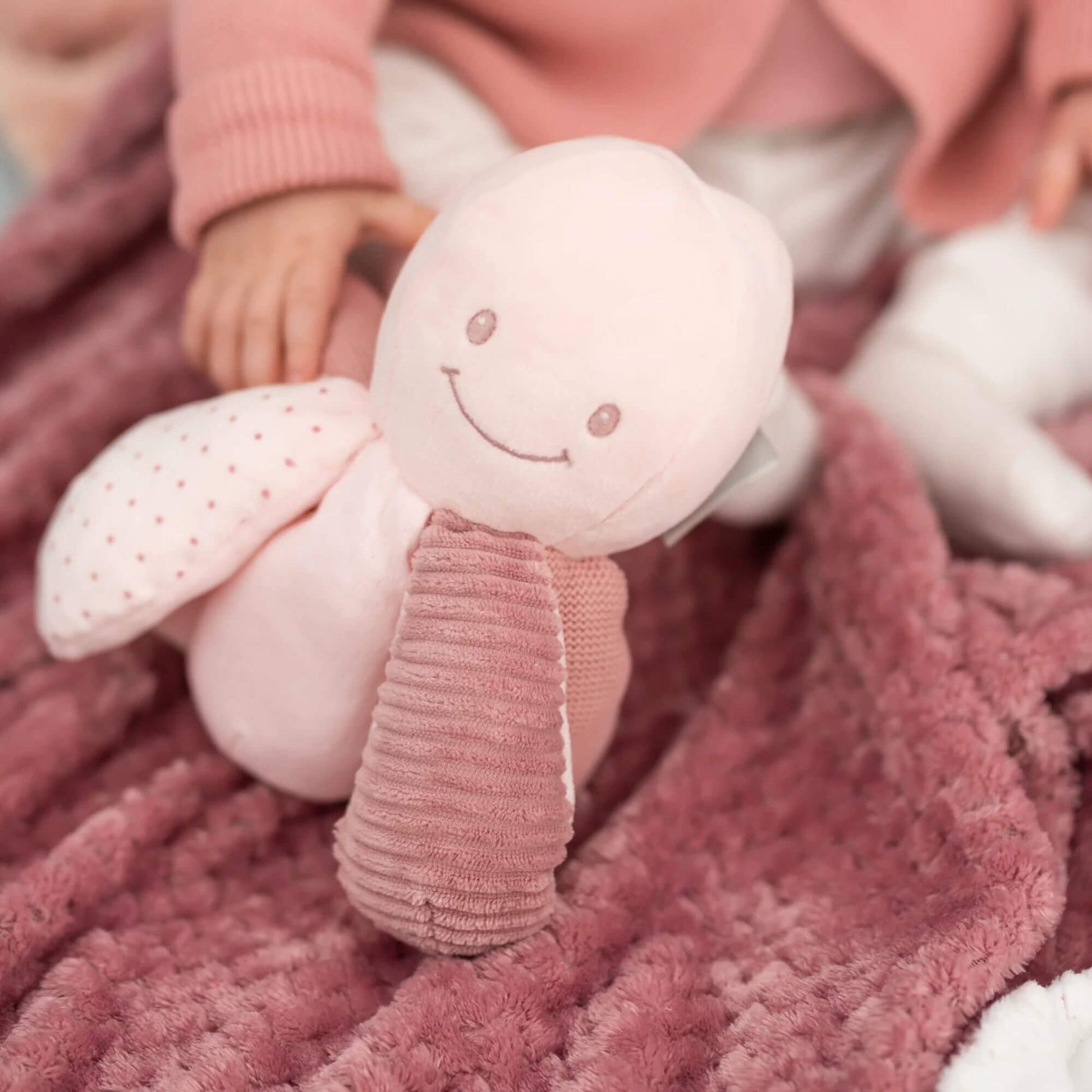 Soft pink Nattou activity octopus with a smiling face, textured tentacles, and spotted accents, lying on a cosy pink blanket.