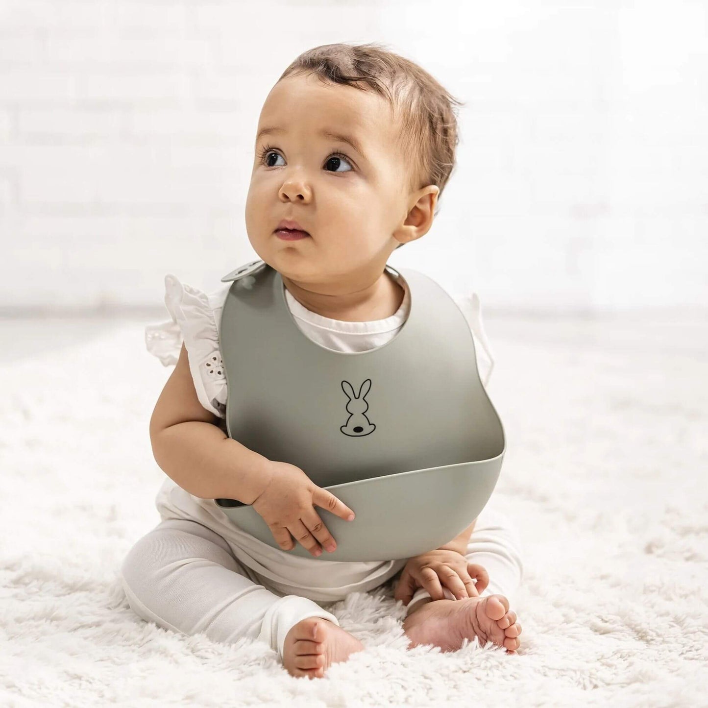 A baby wearing a sage green Nattou silicone bib with an adjustable neck and large food-catching pocket, featuring a cute bunny design, perfect for mess-free feeding.