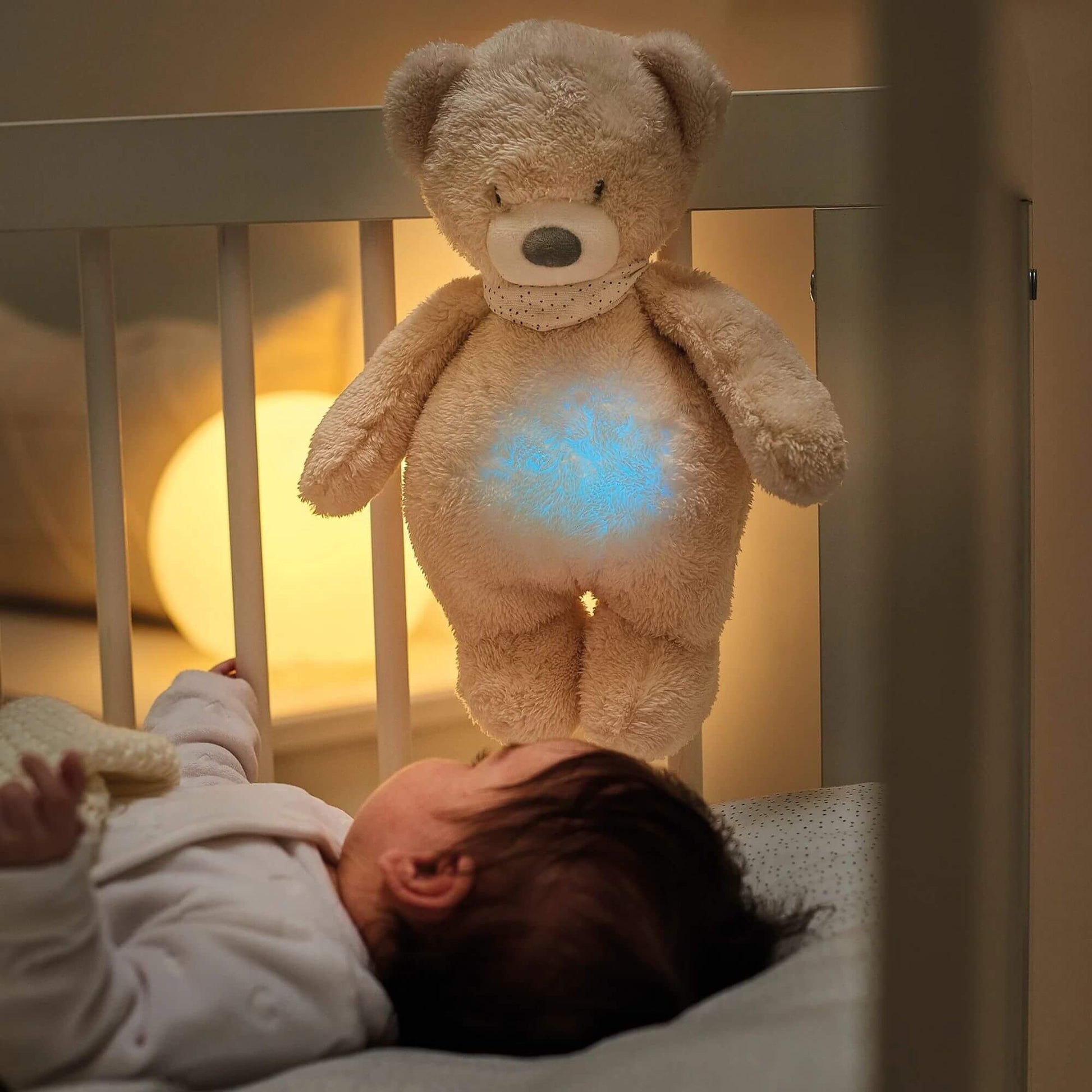 Nattou Sleepy Cuddly bear nightlight hanging on a crib, glowing softly with a blue light as a baby lies in the cot below. Ideal for soothing sleep.